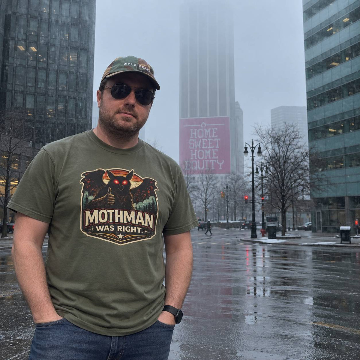 A man wearing an olive green t-shirt with a 'Mothman Was Right' graphic stands on a wet city street in the rain, with skyscrapers and a pink 'Home Sweet Home Chicago' sign in the background.