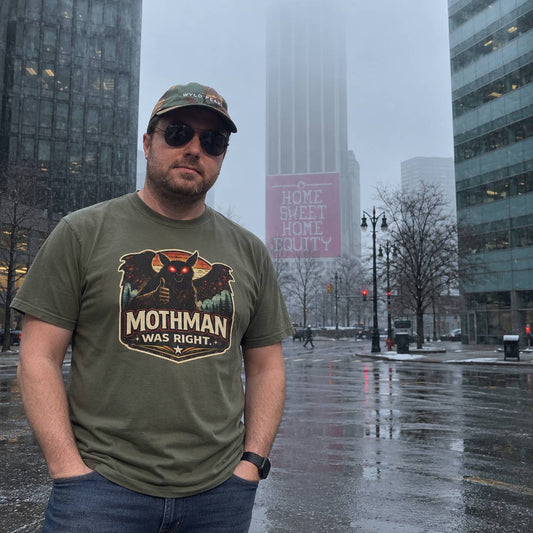A man wearing an olive green t-shirt with a 'Mothman Was Right' graphic stands on a wet city street in the rain, with skyscrapers and a pink 'Home Sweet Home Chicago' sign in the background.