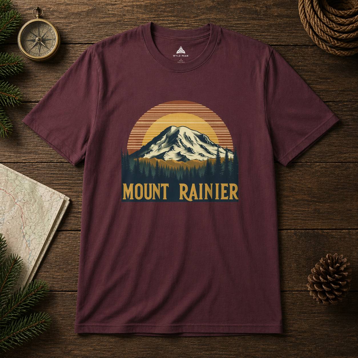 Maroon t-shirt with Mount Rainier graphic on a wooden surface with nature-themed items.
