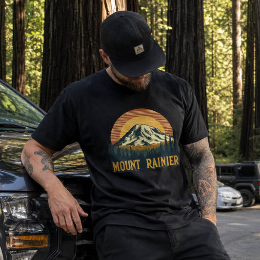 Man wearing a black t-shirt with Mount Rainier design, sitting next to a car in a forest.