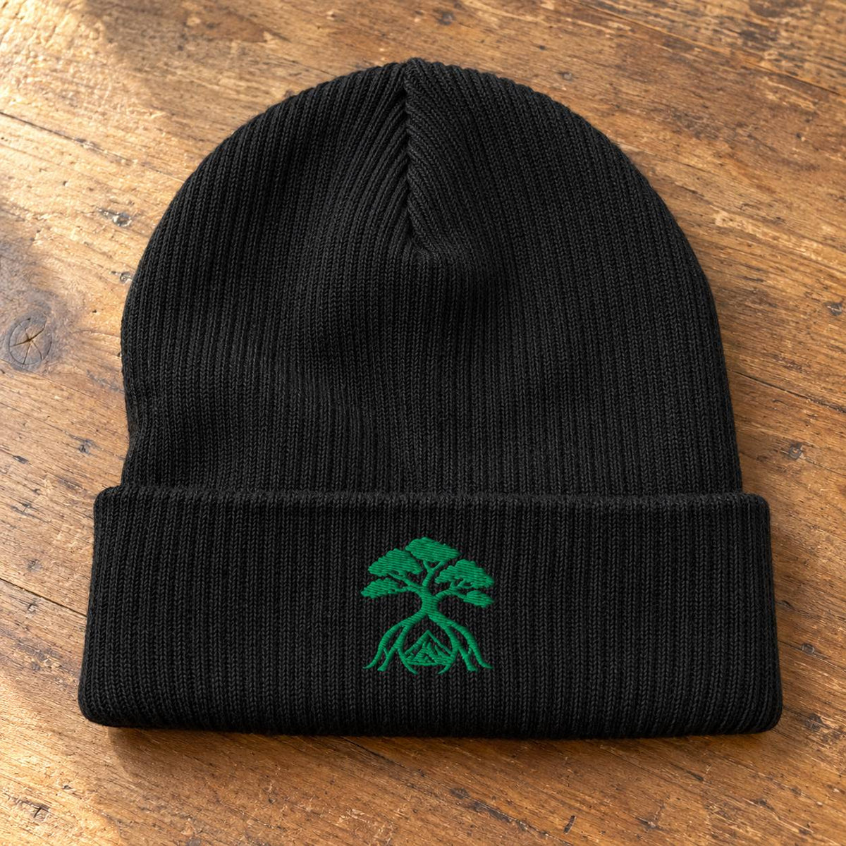 A black ribbed knit beanie hat with a folded cuff, featuring a green embroidered bonsai tree design on the front, displayed on a rustic wooden surface.