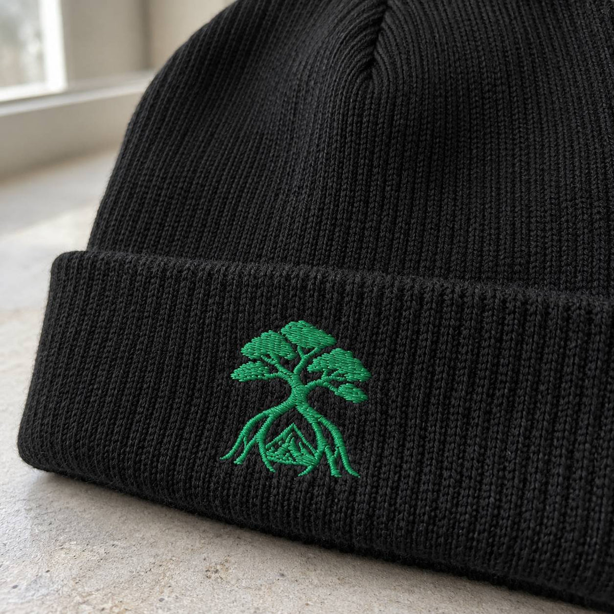 A close-up of a black ribbed knit beanie with a green embroidered bonsai tree and mountain design on the cuff, resting on a light stone surface.