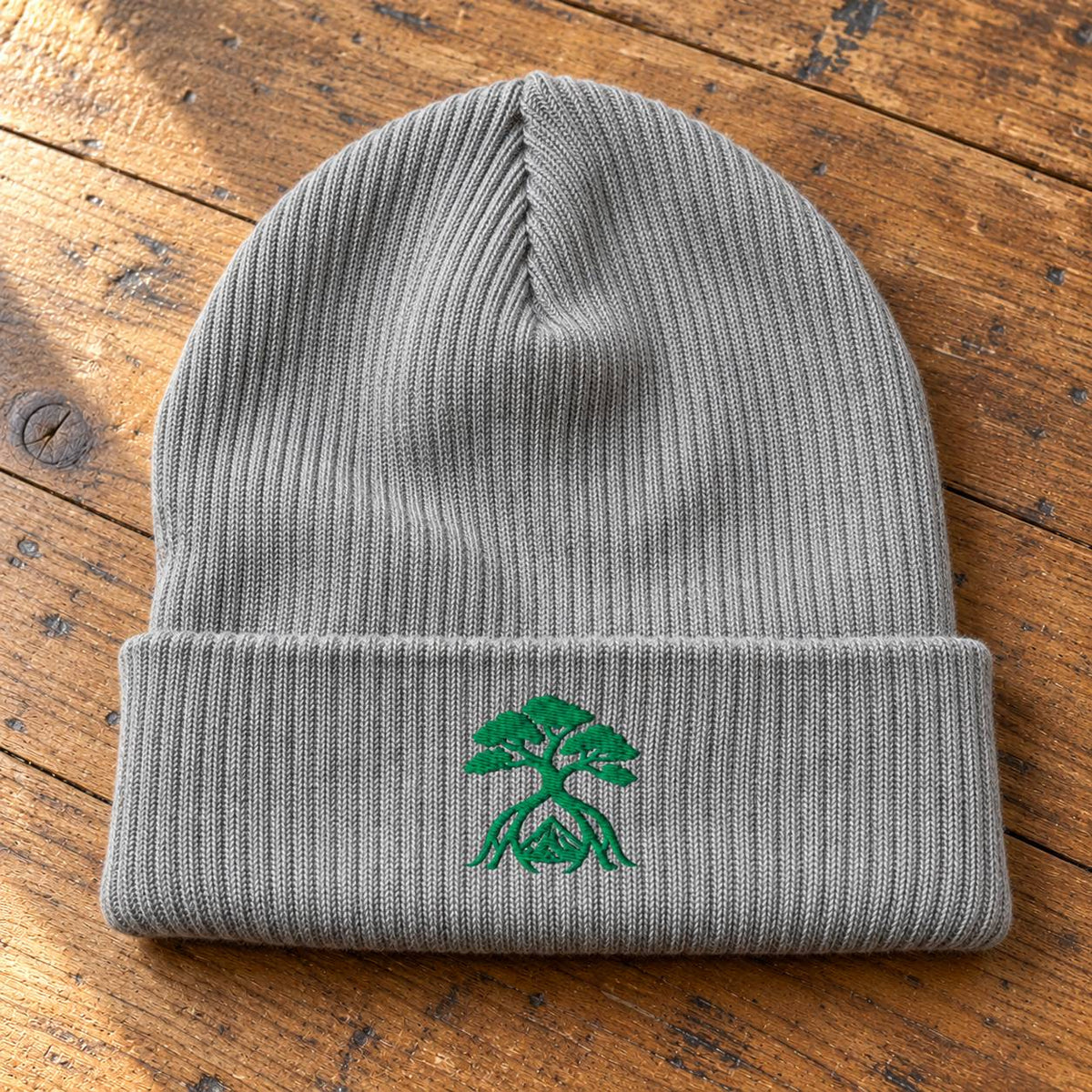A gray ribbed knit beanie hat with a folded cuff, featuring a green embroidered tree-of-life style logo on the front, laid flat on a rustic wooden surface.