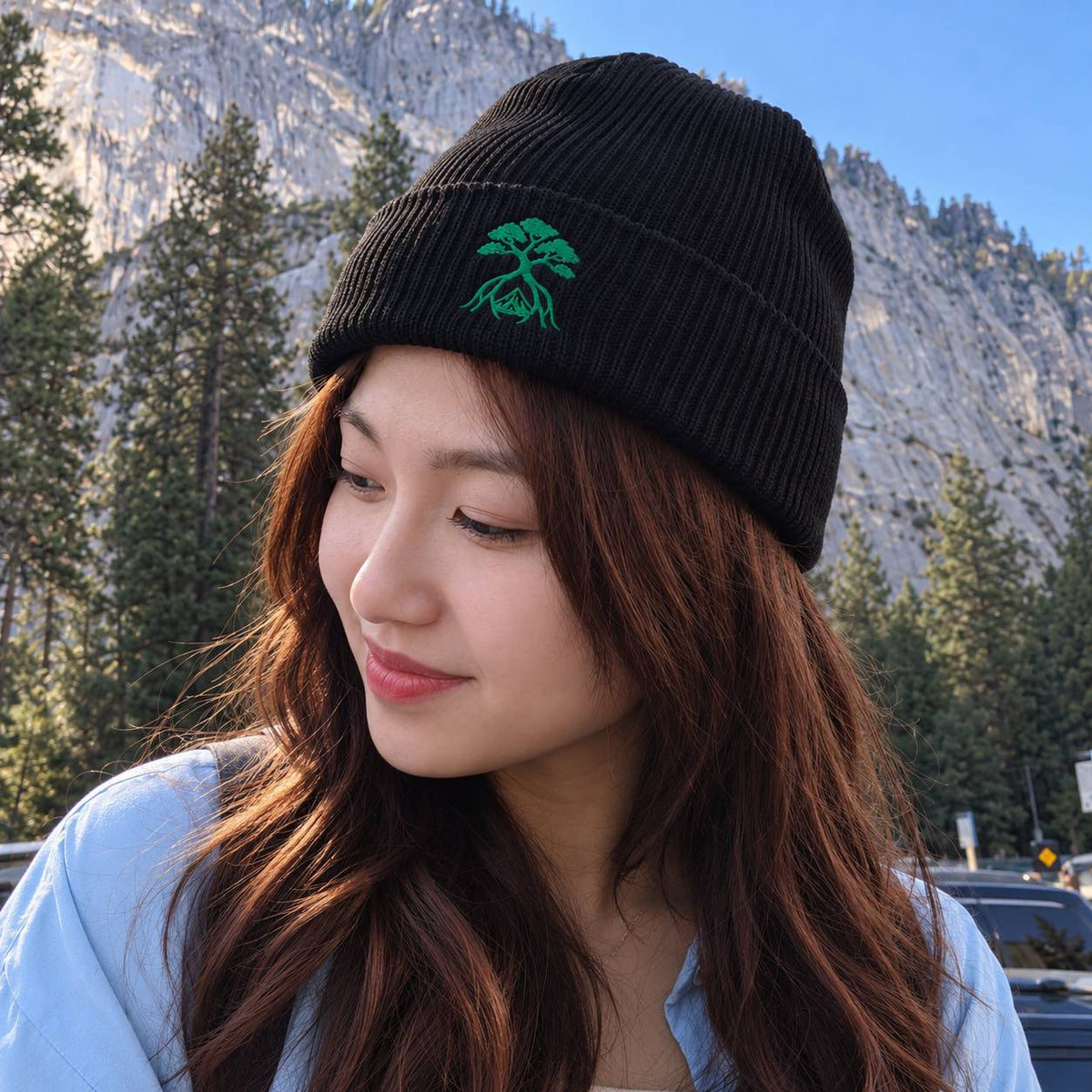 A woman wearing a black ribbed knit beanie with a green embroidered tree logo, photographed outdoors in front of mountains and trees.