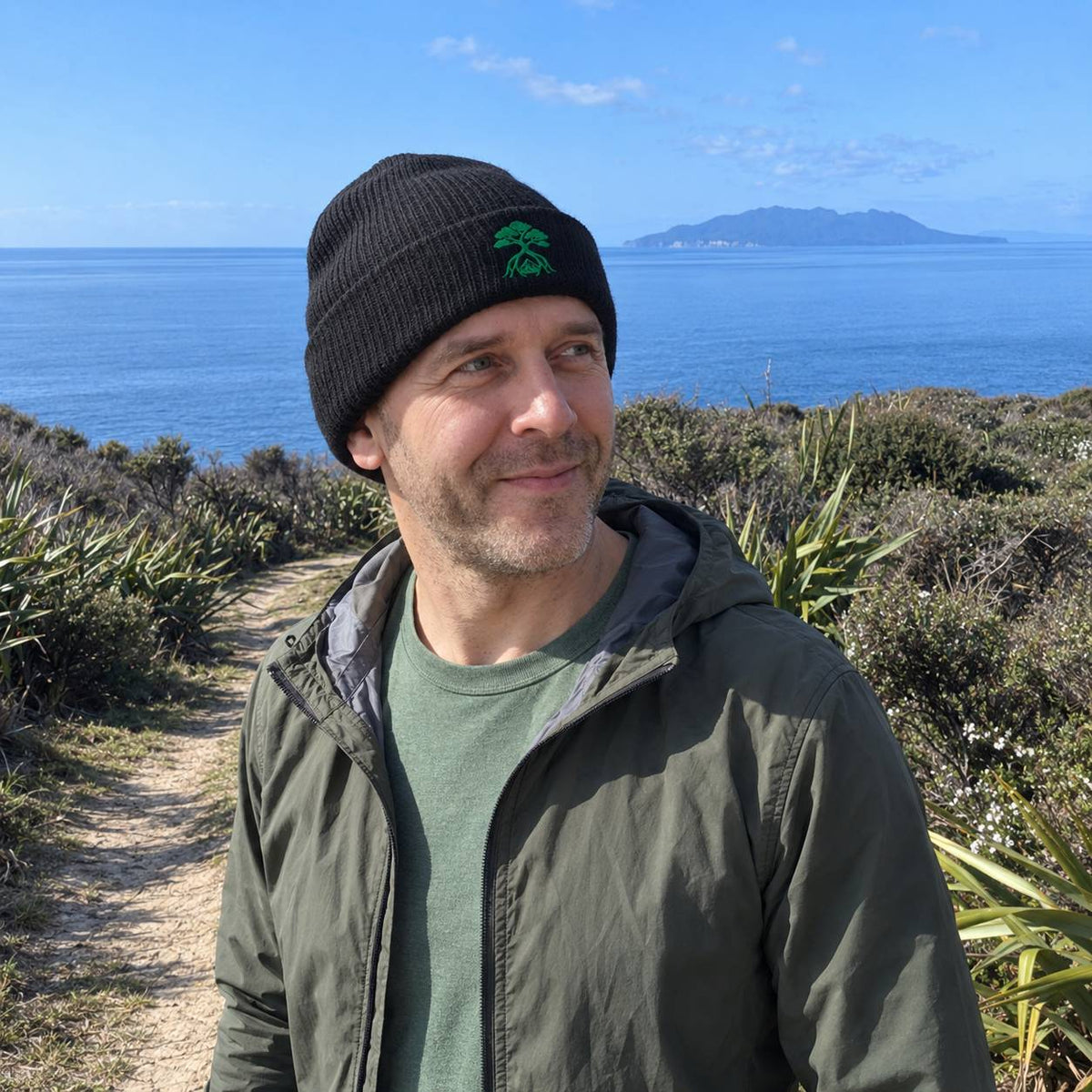A man wearing a black knit beanie with a green tree logo and an olive green jacket over a green t-shirt, standing outdoors on a path with a coastal landscape and ocean in the background.