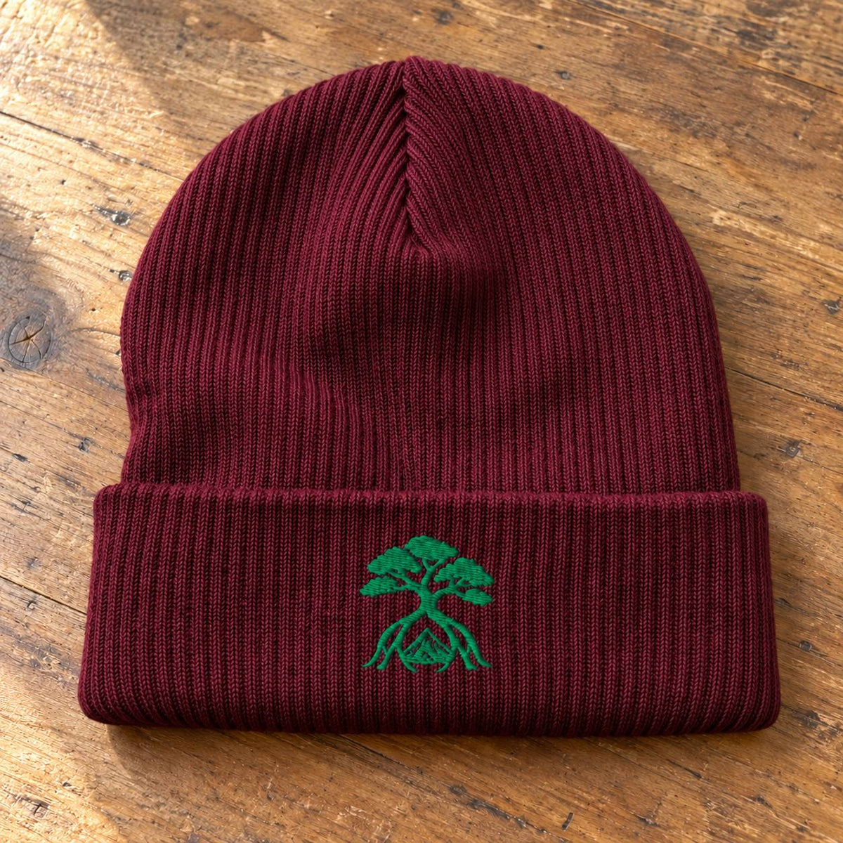 A ribbed knit beanie in deep burgundy with a folded cuff and a small green embroidered bonsai tree logo on the front, displayed flat on a rustic wooden surface.