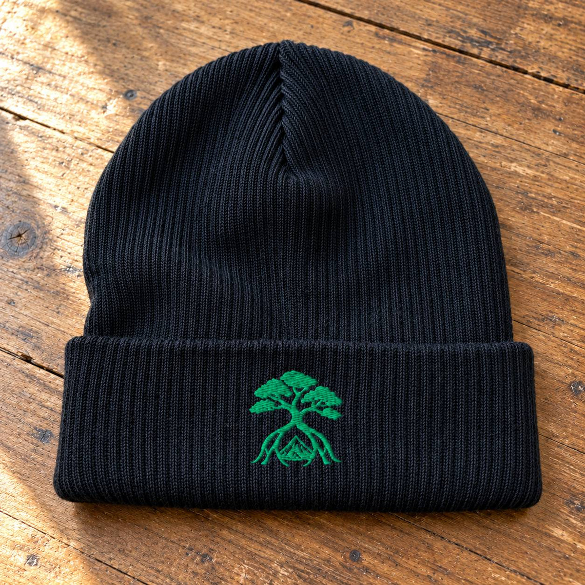 A black ribbed knit beanie with a folded cuff, featuring an embroidered green bonsai tree logo on the front, displayed on a rustic wooden surface.