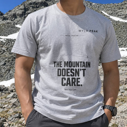 A man wearing a light gray heather crewneck t-shirt with black text that reads 'THE MOUNTAIN DOESN'T CARE.' and 'WYLD PEAK' on the chest, set against a rocky mountain background with patches of snow.