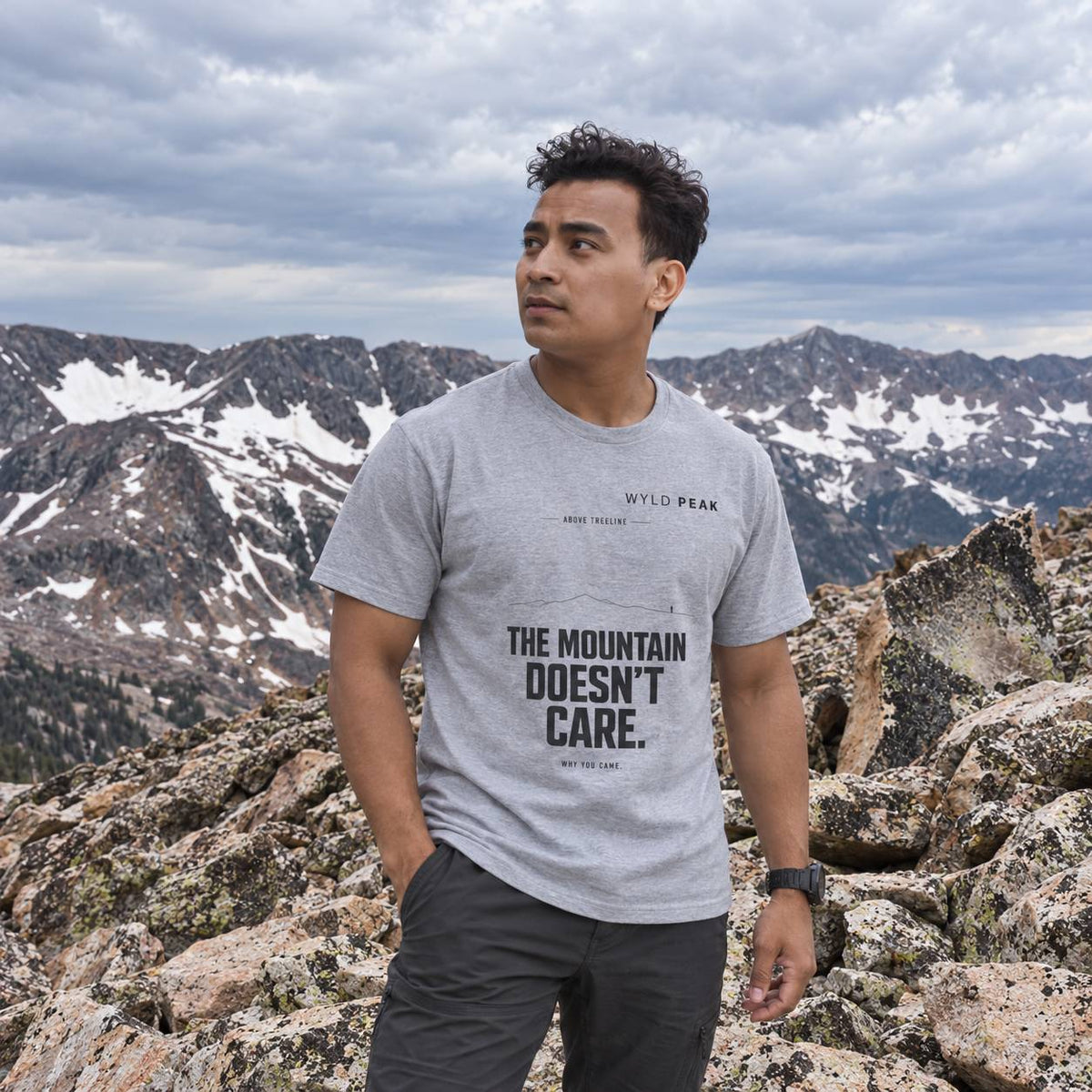 A man wearing a light gray t-shirt with the text 'THE MOUNTAIN DOESN'T CARE.' stands on a rocky mountain peak with snowy peaks and a cloudy sky in the background.