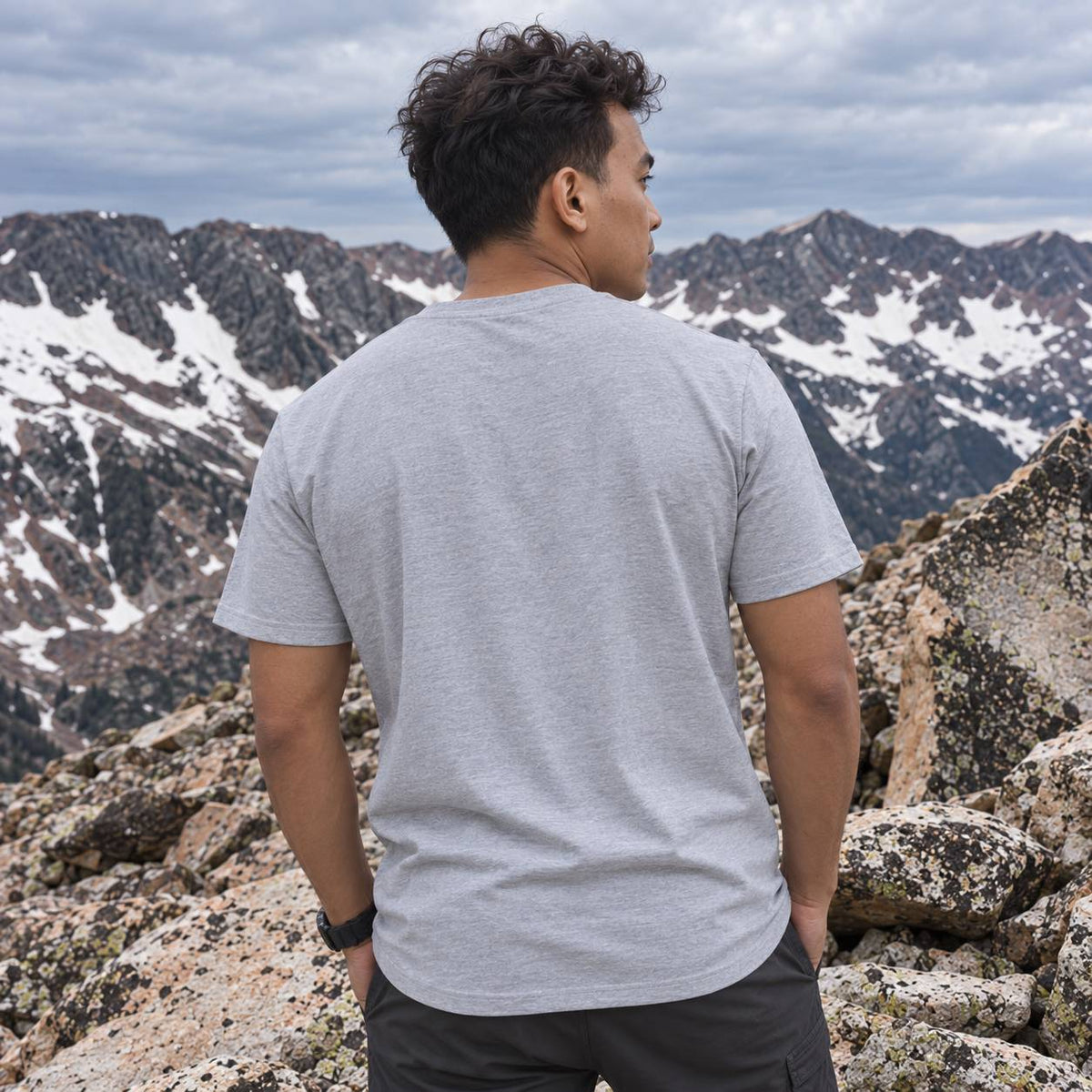 A man wearing a light gray heather crewneck t-shirt and dark gray pants, standing on a rocky mountain peak with snow-capped mountains in the background under a cloudy sky.