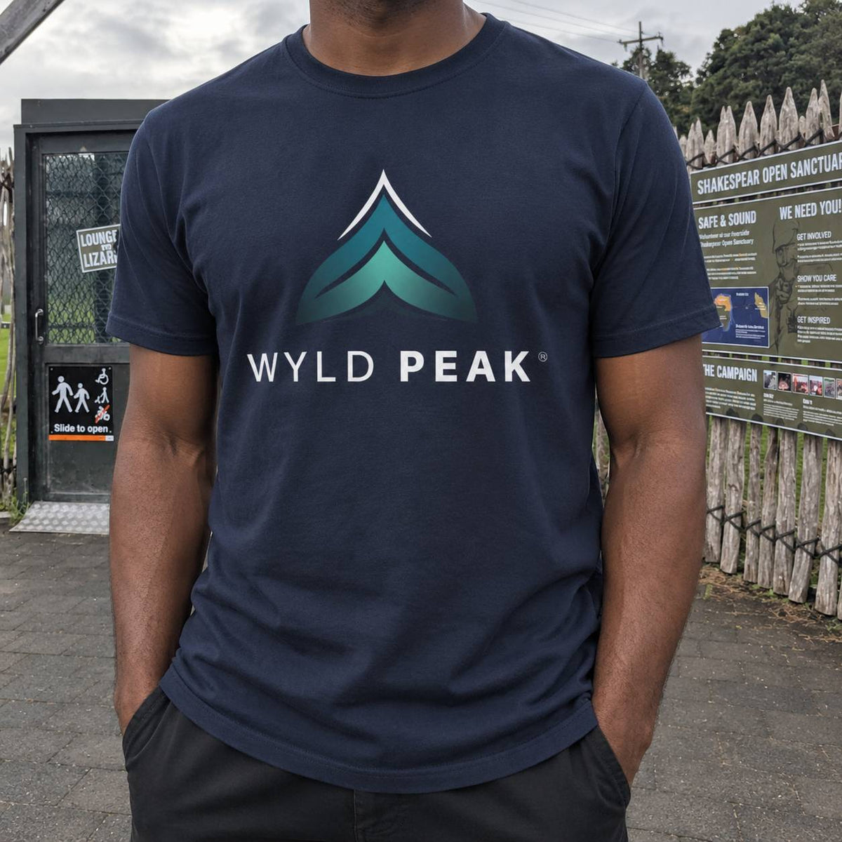 A man wearing a navy blue t-shirt with a 'WYLD PEAK' graphic printed on the chest, standing outdoors in front of a fence and sign.