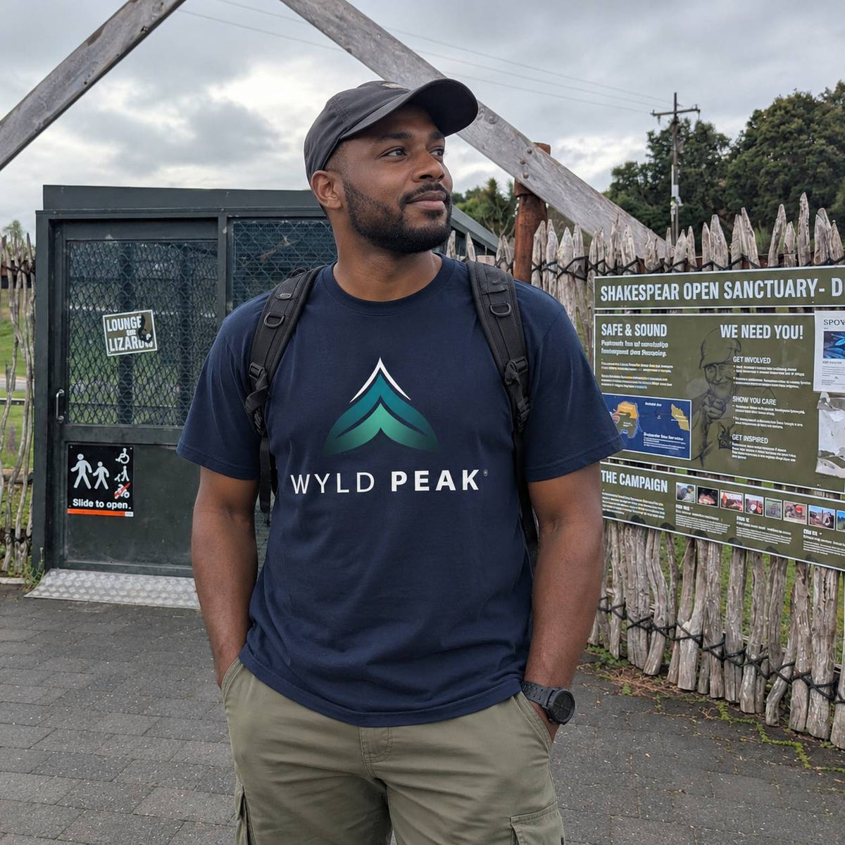 A man wearing a navy blue t-shirt with a green and blue mountain logo and the text 'WYLD PEAK' stands outdoors in front of a gate and informational sign.