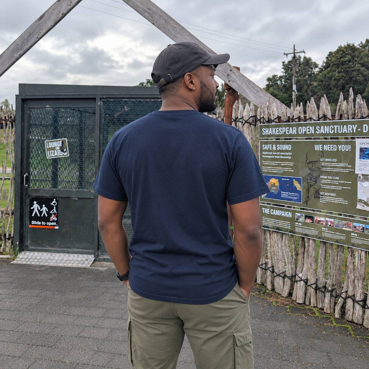 A man wearing a navy blue t-shirt and khaki cargo pants stands with his back to the camera, looking at a sign at an outdoor sanctuary. The sign reads 'Shakespeare Open Sanctuary - D'.