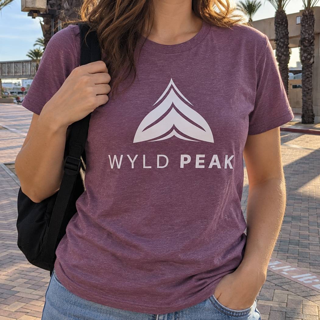 A woman wearing a heathered purple t-shirt with a white graphic logo that reads 'WYLD PEAK' and a mountain peak icon, photographed outdoors on a sunny day.