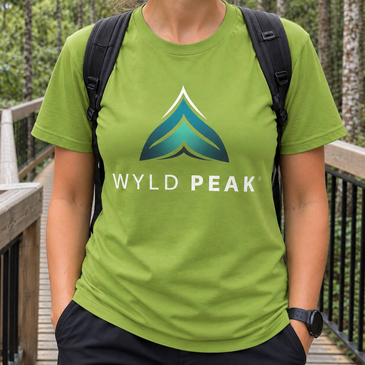 A person wearing a bright green crewneck t-shirt with a 'WYLD PEAK' logo printed on the chest, standing on a wooden boardwalk outdoors.
