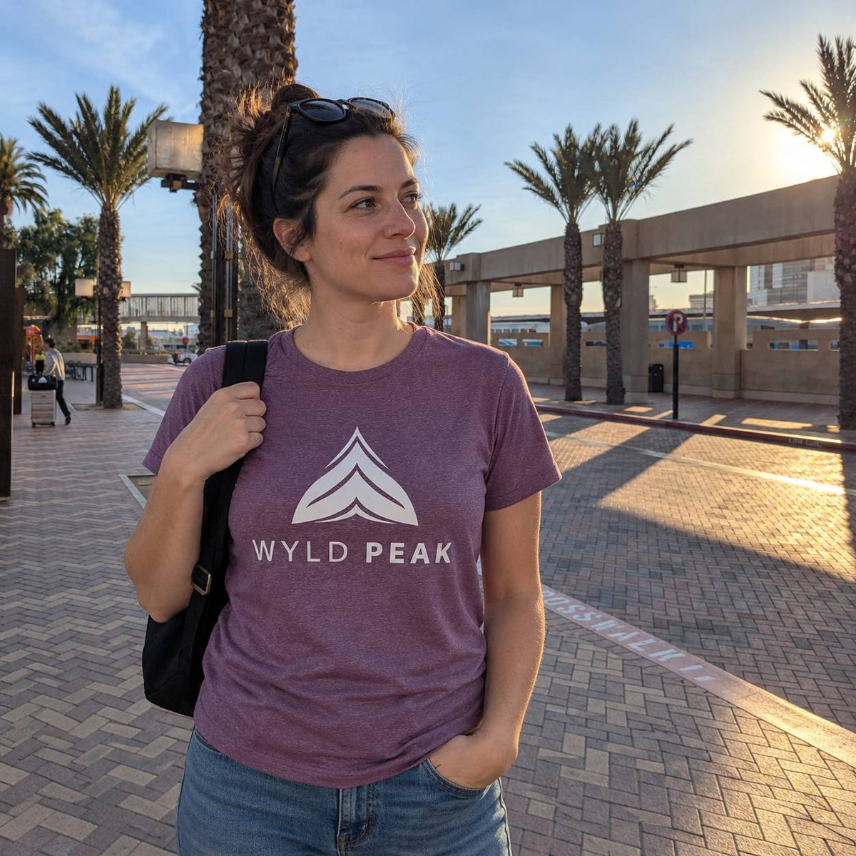 A woman wearing a heathered purple t-shirt with a white 'WYLD PEAK' logo and text, standing outdoors on a paved walkway with palm trees and a building in the background.