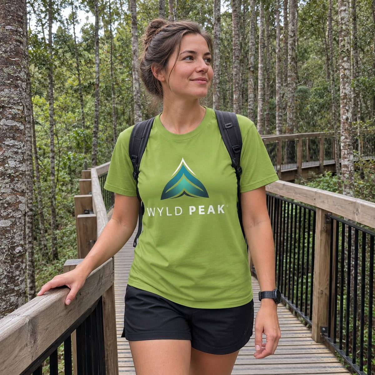 A woman wearing a green t-shirt with a 'WYLD PEAK' logo stands on a wooden boardwalk surrounded by a forest.