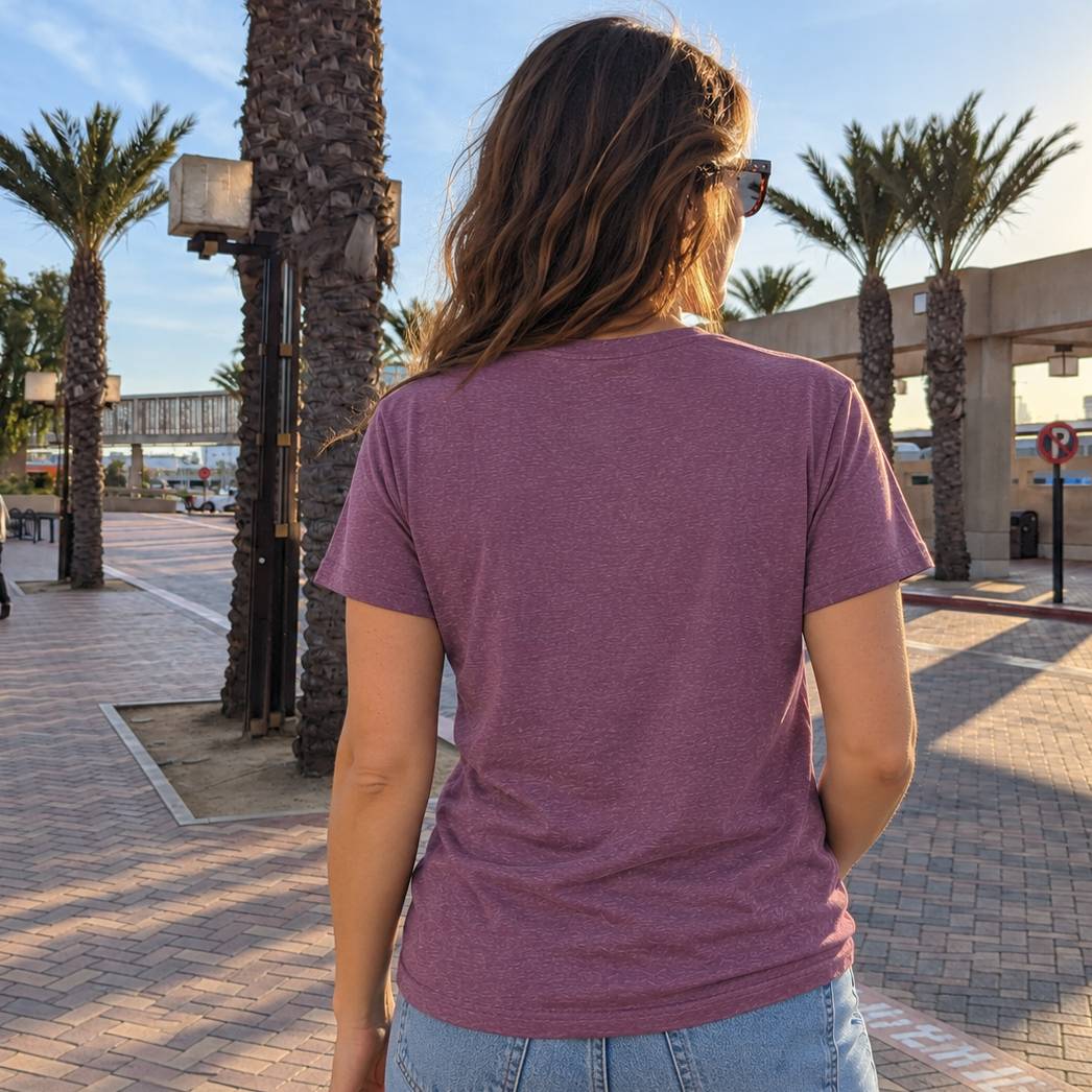 A woman viewed from behind wearing a heathered mauve-purple short-sleeve t-shirt and light blue jeans, standing outdoors on a paved walkway with palm trees and a building in the background.