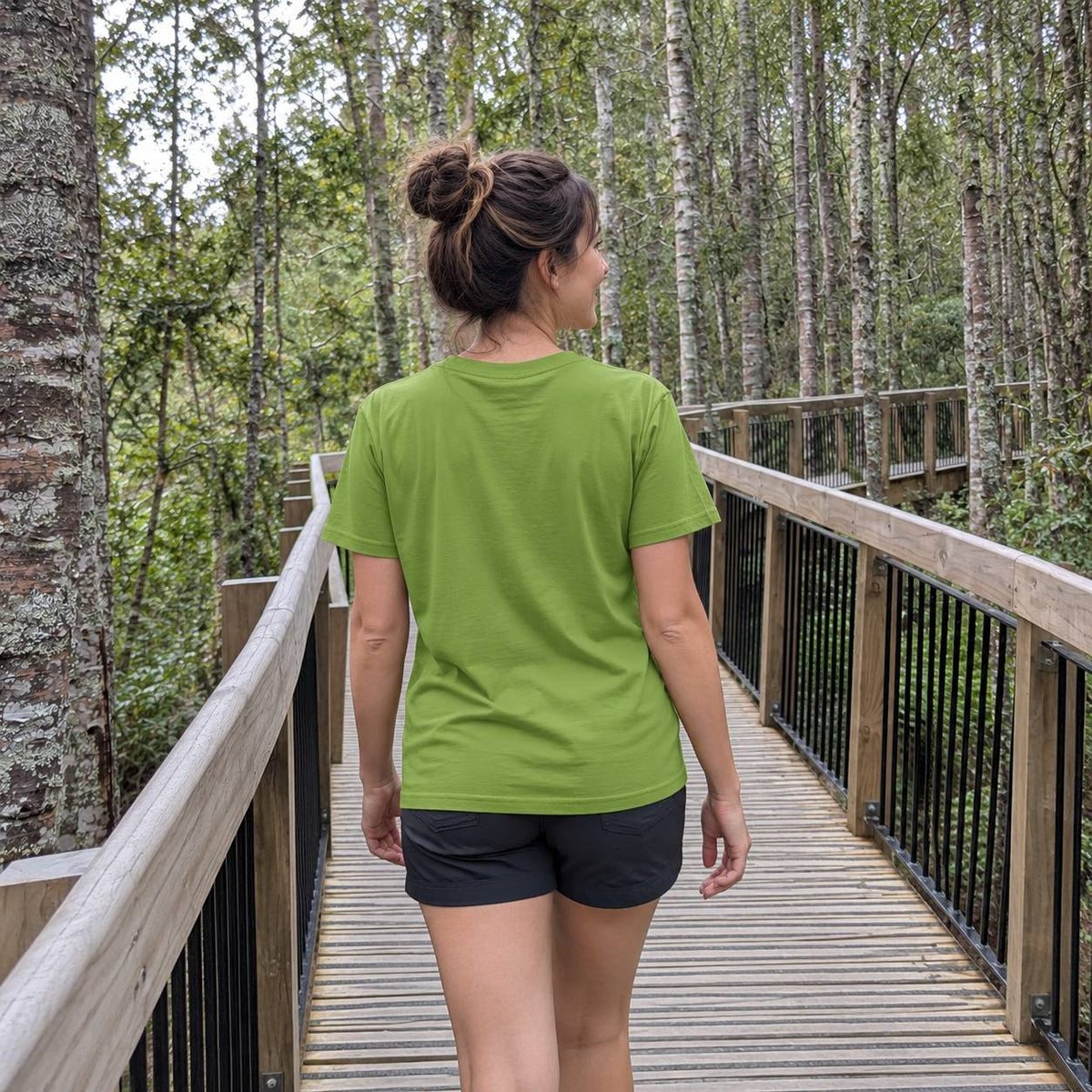 A woman viewed from behind wearing a lime green short-sleeve t-shirt and black athletic shorts, walking on a wooden boardwalk surrounded by a dense forest.