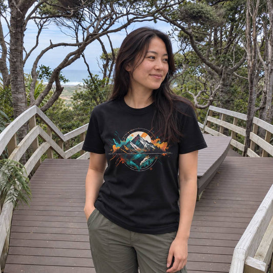 A woman wearing a black graphic t-shirt and olive green pants stands on a wooden boardwalk surrounded by trees, looking to the side. The t-shirt features a colorful graphic of a mountain and water in shades of teal, orange, and white.