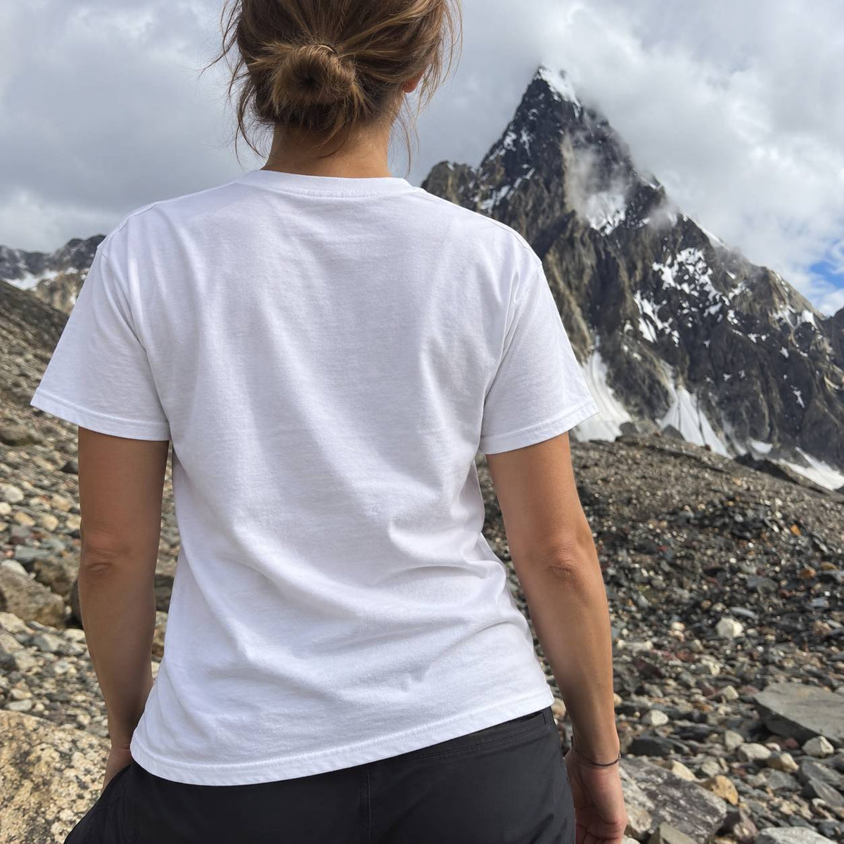 A person viewed from behind wearing a plain white short-sleeve T-shirt and dark shorts, standing on a rocky mountainside with a snow-dusted peak in the background.