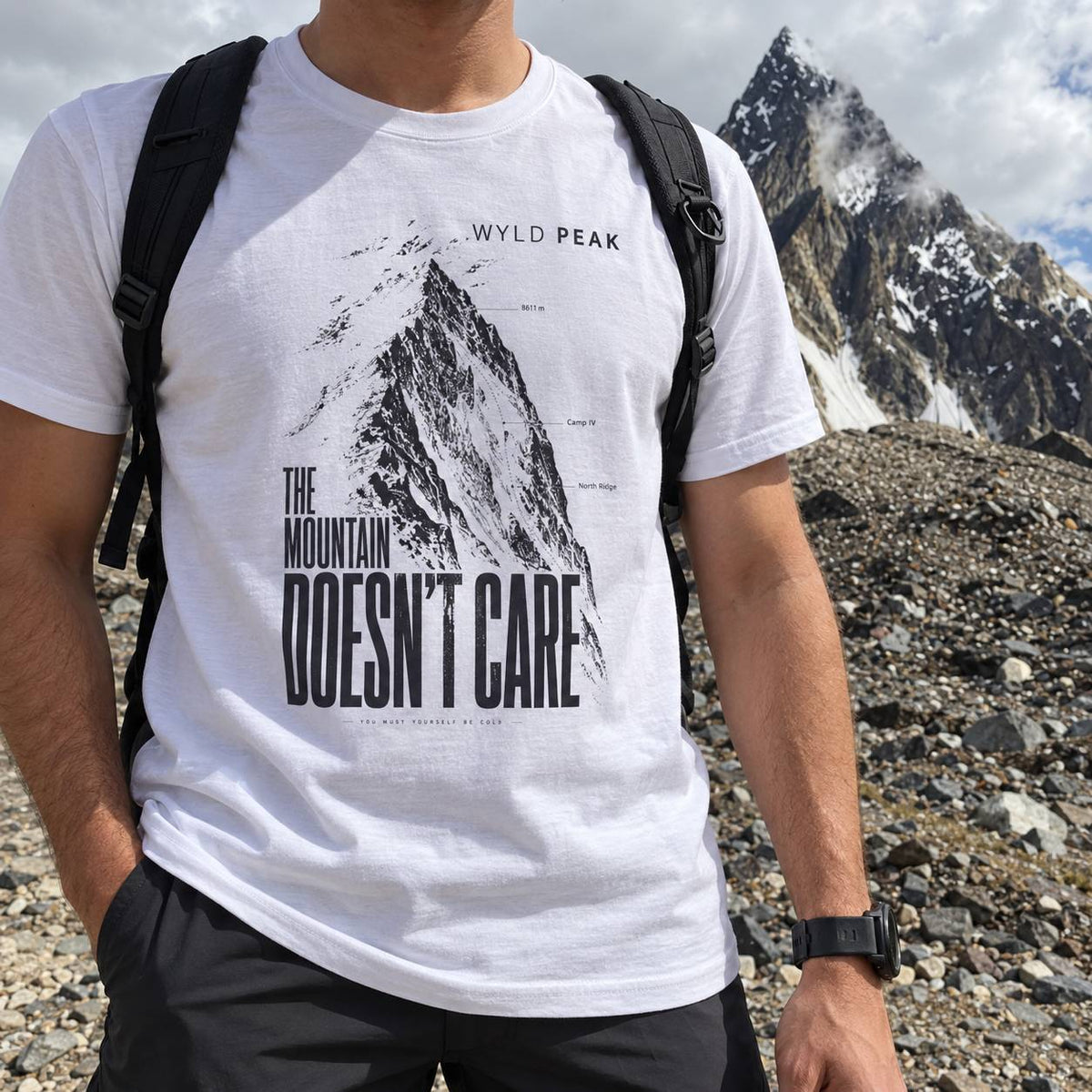 A man wearing a white graphic t-shirt with a black print of a mountain and the text 'THE MOUNTAIN DOESN'T CARE' and 'WYLD PEAK'. He is outdoors in a rocky mountain landscape with a snow-capped peak in the background.