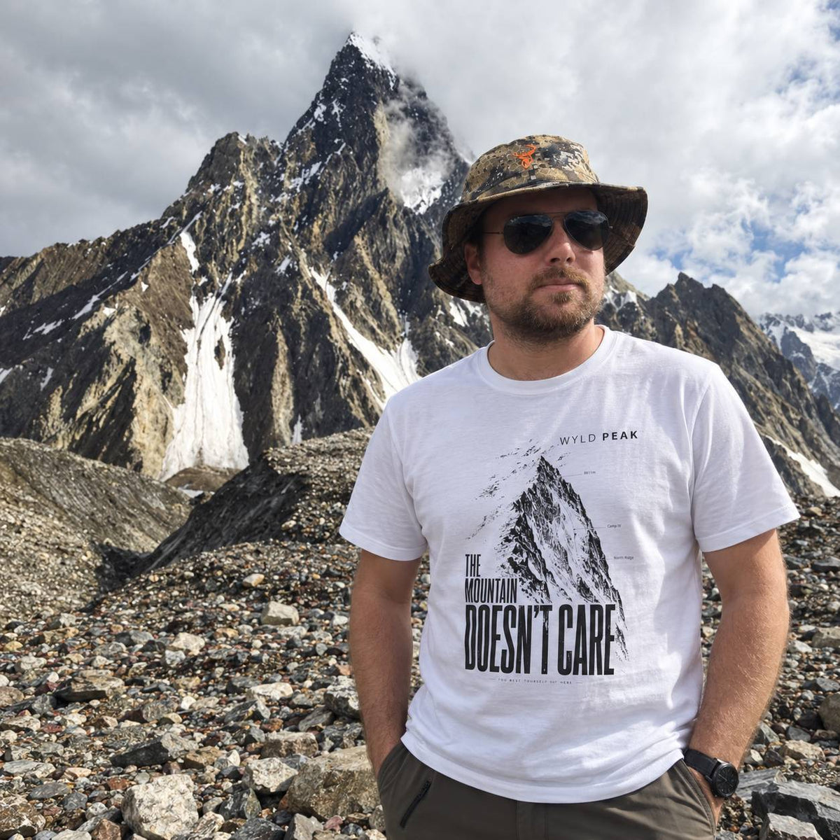 A man wearing a white graphic t-shirt with 'THE MOUNTAIN DOESN'T CARE' and 'WYLD PEAK' printed on the front, standing in a rocky mountain landscape with a sharp peak in the background.
