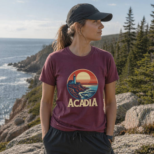 Person wearing a 'Acadia' t-shirt with a lighthouse design, standing on a rocky landscape.