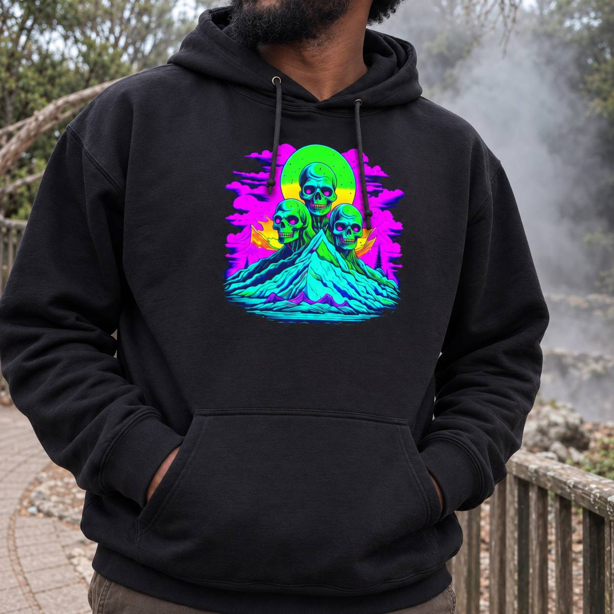 A man wearing a black pullover hoodie with a vibrant, colorful graphic print on the chest showing three skulls on a mountain peak with neon green, pink, and yellow hues. The background is an outdoor setting with trees and a railing.