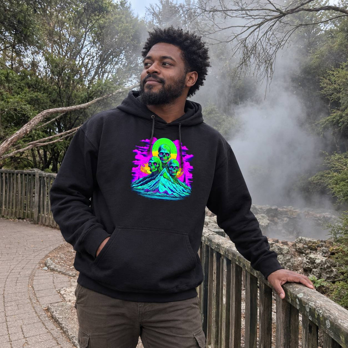 A man wearing a black hoodie with a colorful neon-style graphic print of a figure in a green and purple landscape, standing outdoors on a wooden boardwalk with trees and steam in the background.
