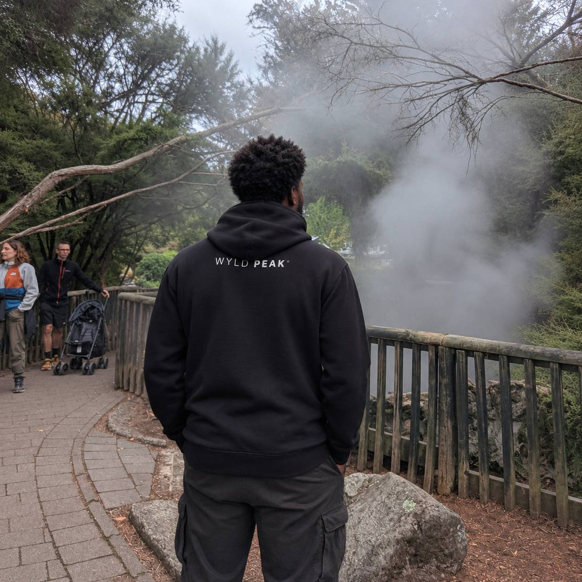 A man viewed from behind wearing a black pullover hoodie with 'WYLD PEAK' printed in white on the back, standing on a paved path in a misty outdoor park setting.
