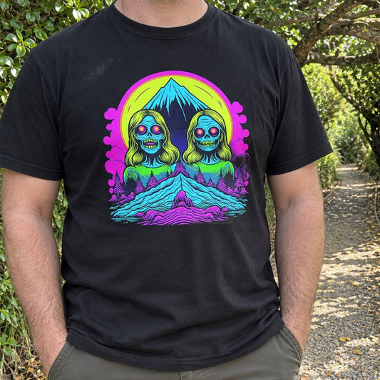 A man wearing a black t-shirt with a vibrant, colorful retro-style graphic print of two skull-faced figures in front of a mountain. The background is an outdoor path with trees.