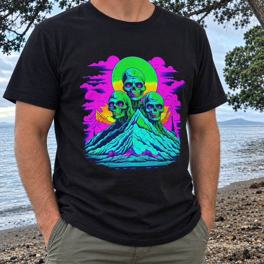 A man wearing a black t-shirt with a vibrant neon green, pink, and blue graphic of three skulls on a mountain peak, set against a beach background.