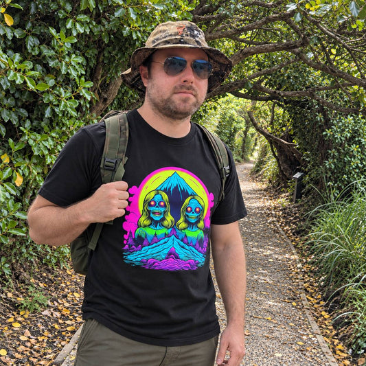 A man wearing a black t-shirt with a colorful psychedelic graphic of two mirrored figures in front of a mountain, standing on a wooded path.