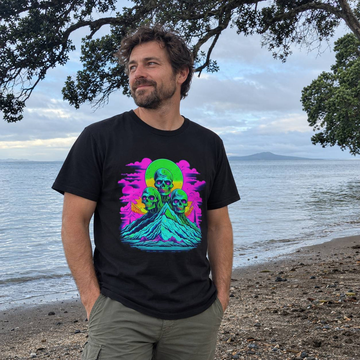 A man standing on a beach wearing a black t-shirt with a colorful graphic print of three skulls on a mountain, with the ocean and trees in the background.