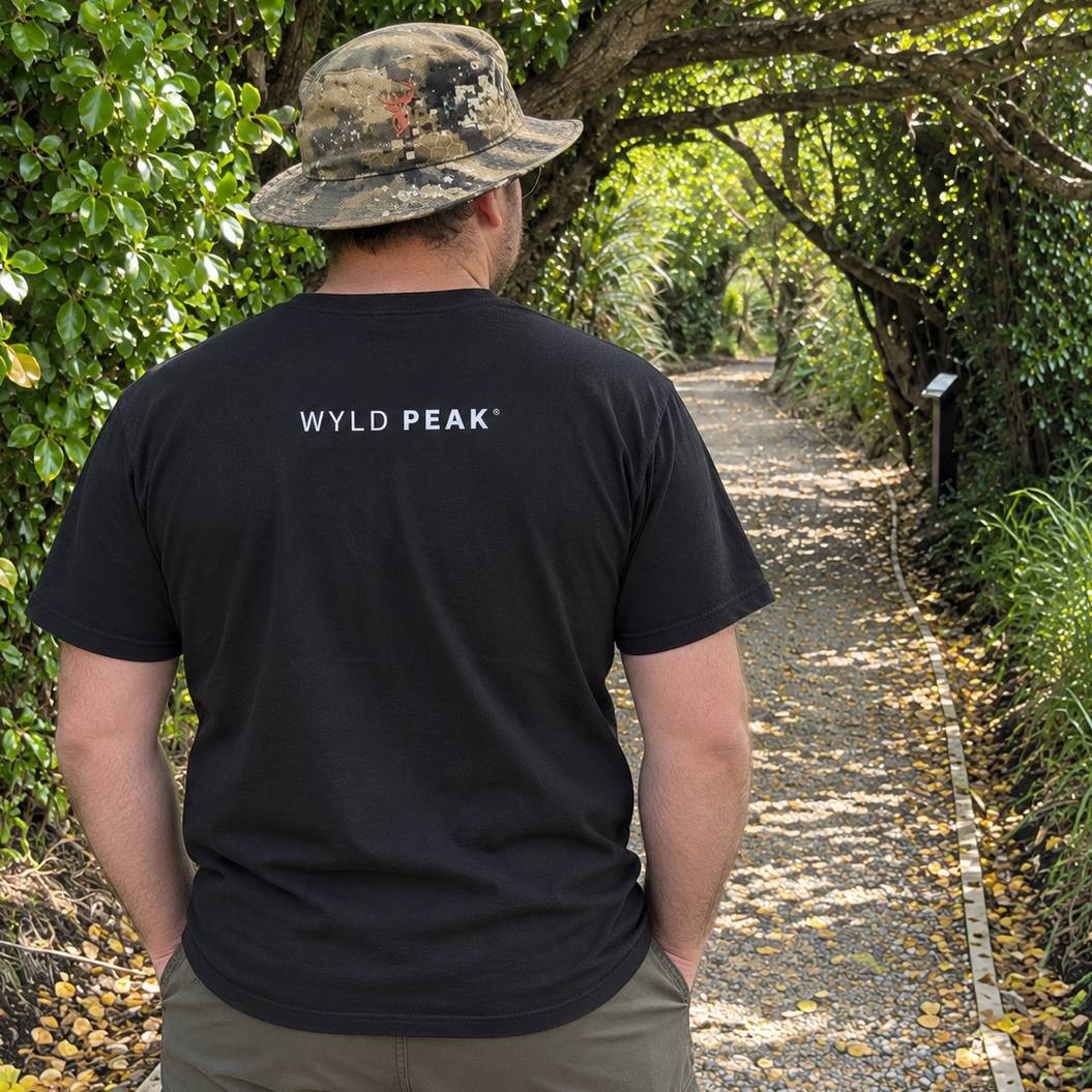 A man viewed from behind wearing a black t-shirt with 'WYLD PEAK' printed in white on the back, walking on a leaf-covered path surrounded by green trees. He is also wearing a camouflage bucket hat.