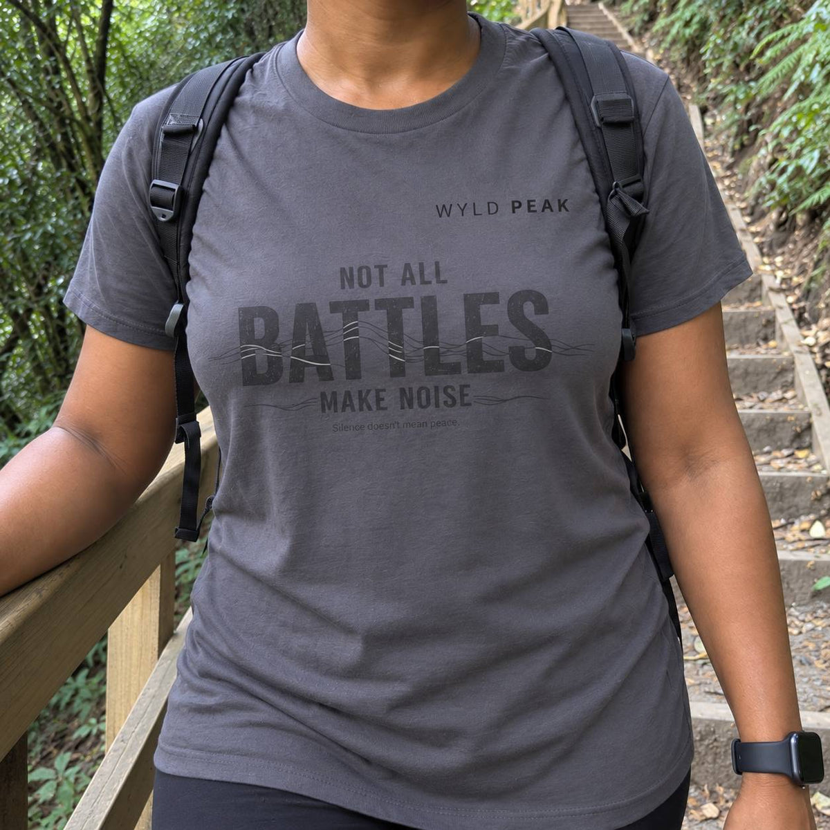 A person wearing a dark gray heather graphic t-shirt with the text 'WYLD PEAK NOT ALL BATTLES MAKE NOISE' in a forest setting on a wooden path.