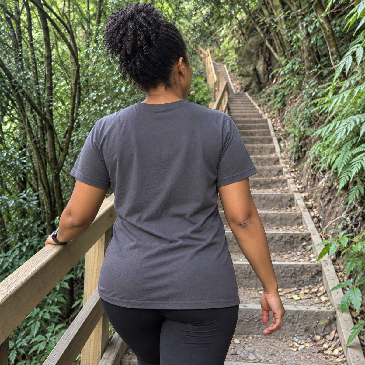 A woman viewed from behind wearing a dark gray crewneck t-shirt and black leggings, walking up a stone staircase in a lush green forest.