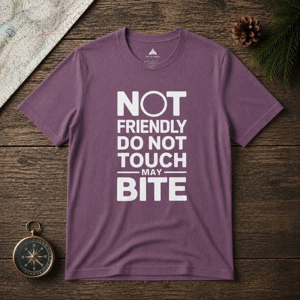 Purple t-shirt with white text 'Not friendly do not touch may bite' on a wooden surface with a compass and pine cone.