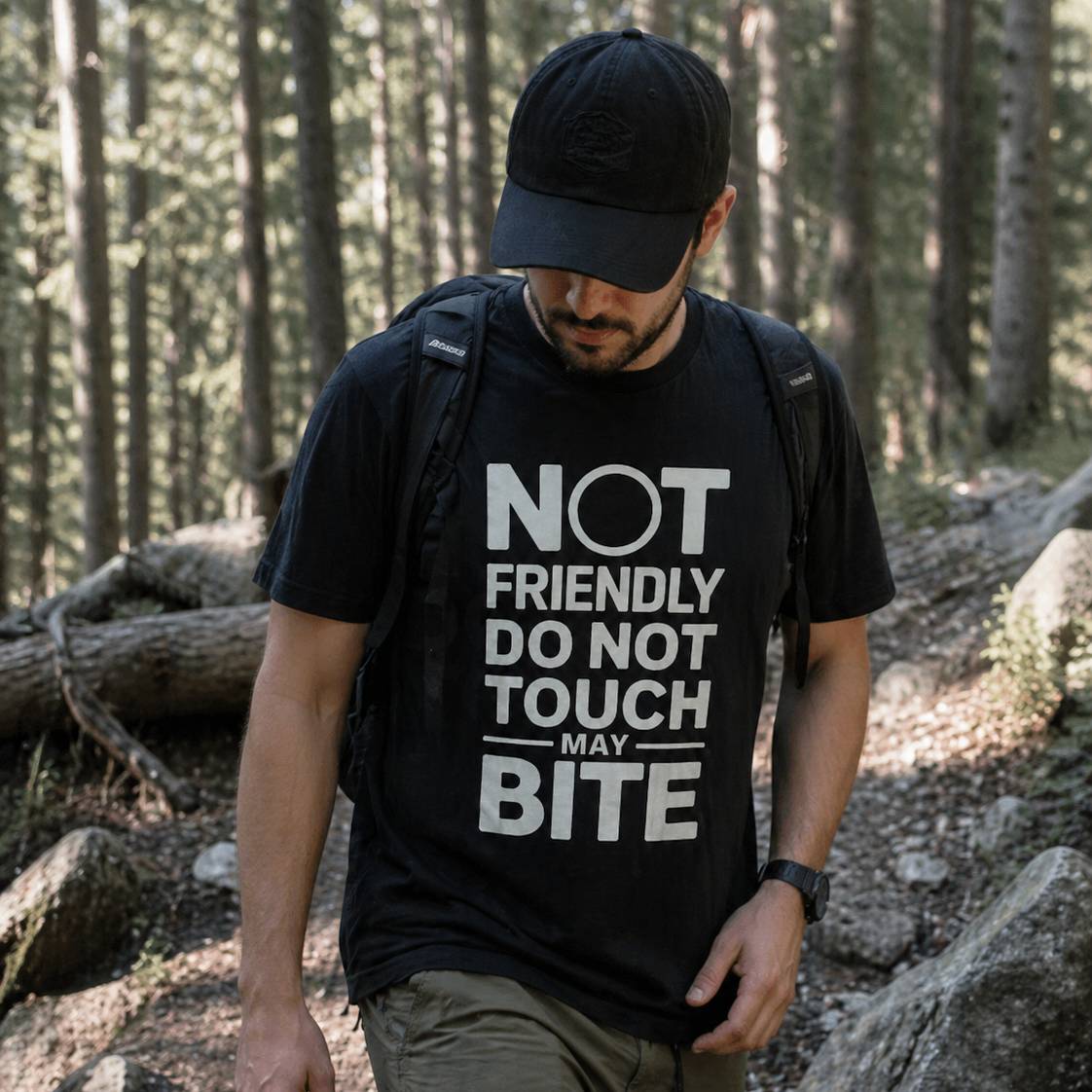 Man hiking in a forest wearing a black t-shirt with text and a black cap.