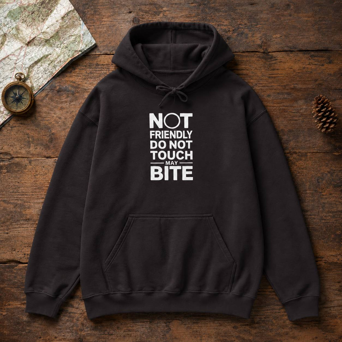 A black pullover hoodie with a white graphic text print reading 'NOT FRIENDLY DO NOT TOUCH MAY BITE' laid flat on a rustic wooden surface with a map, compass, and pinecone as props.