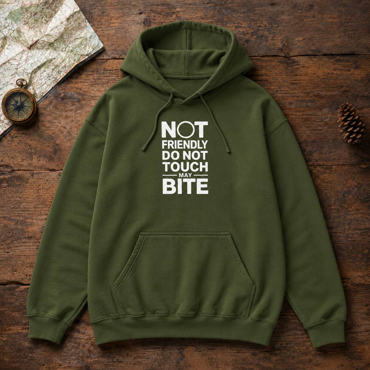 An olive green pullover hoodie with a white graphic text print reading 'NOT FRIENDLY DO NOT TOUCH MAY BITE' laid flat on a rustic wooden surface with a map, compass, and pinecone as props.