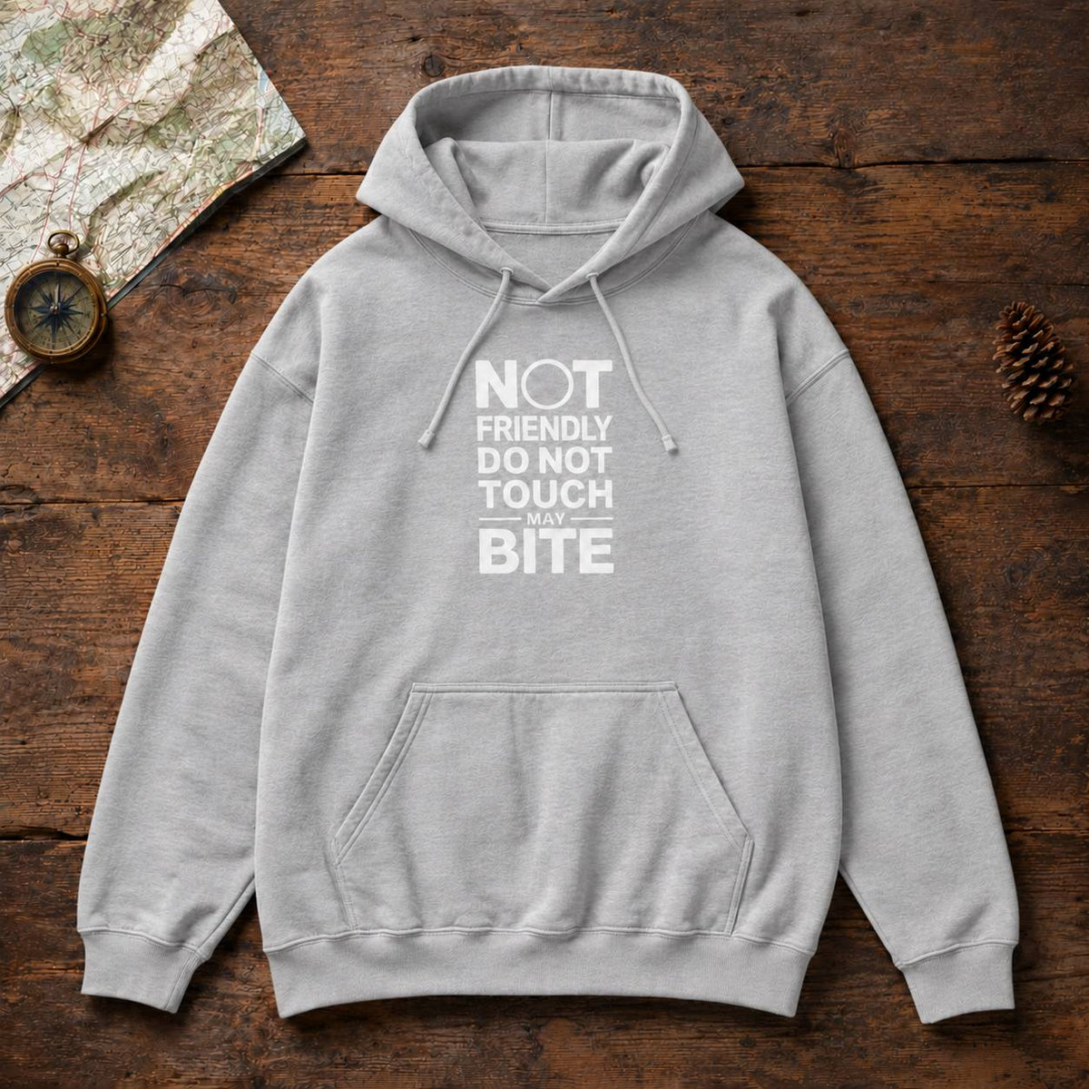 A light gray pullover hoodie with a white graphic text print reading 'NOT FRIENDLY DO NOT TOUCH MAY BITE' laid flat on a rustic dark wood background with a map, compass, and pinecone as props.