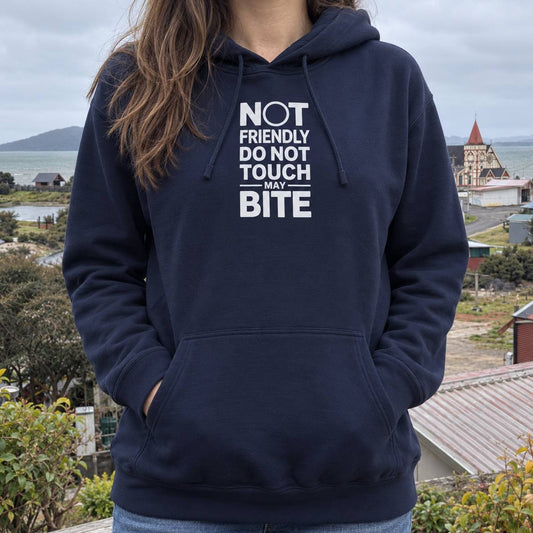 A woman wearing a navy blue hoodie with a white graphic text print that reads 'NOT FRIENDLY DO NOT TOUCH MAY BITE'. The background shows an outdoor coastal landscape with houses, water, and a cloudy sky.