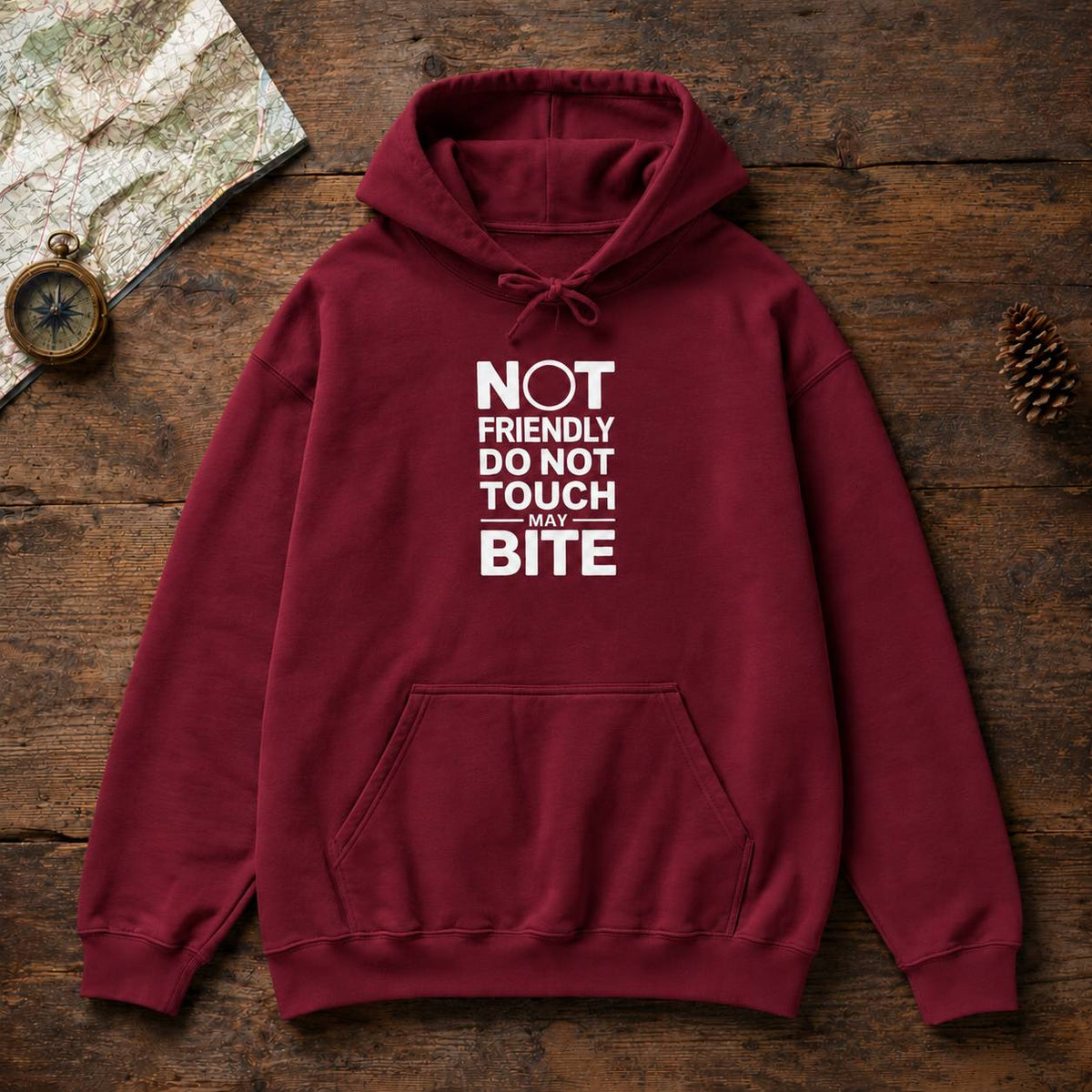 A burgundy pullover hoodie with a white graphic text print reading 'NOT FRIENDLY DO NOT TOUCH MAY BITE' laid flat on a rustic wooden surface with a map, compass, and pinecone props.