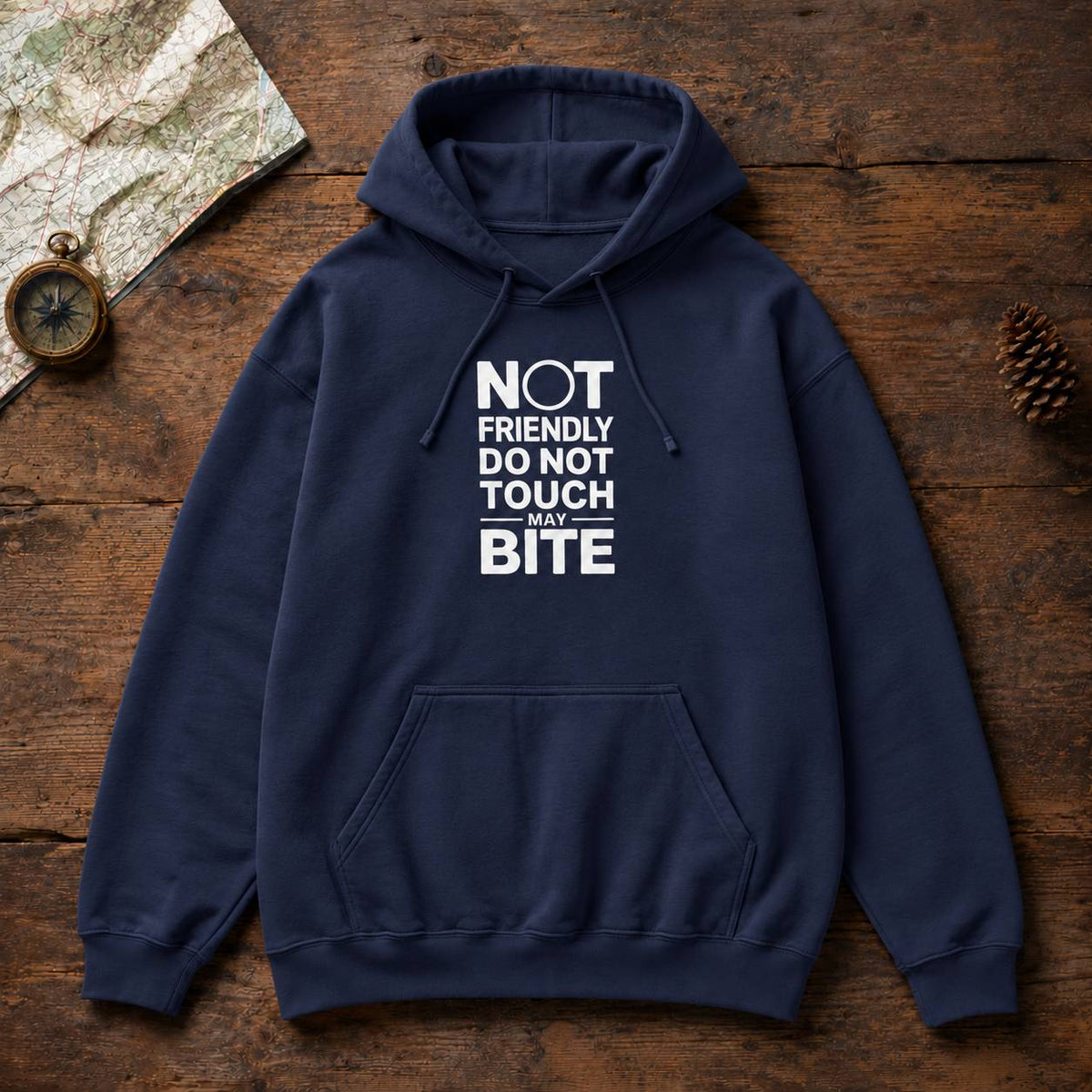 A navy blue pullover hoodie with a white graphic text print reading 'NOT FRIENDLY DO NOT TOUCH MAY BITE' laid flat on a rustic wooden surface with a map, compass, and pinecone as props.