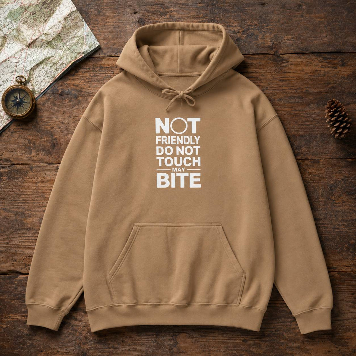 A tan pullover hoodie laid flat on a rustic wooden surface, with a white printed slogan reading 'NOT FRIENDLY DO NOT TOUCH MAY BITE' on the chest. A front kangaroo pocket and drawstring hood are visible.