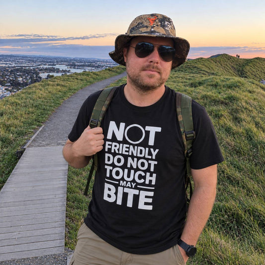 A man wearing a black graphic t-shirt with the text 'NOT FRIENDLY DO NOT TOUCH MAY BITE' in white, standing on a grassy hilltop path at sunset.