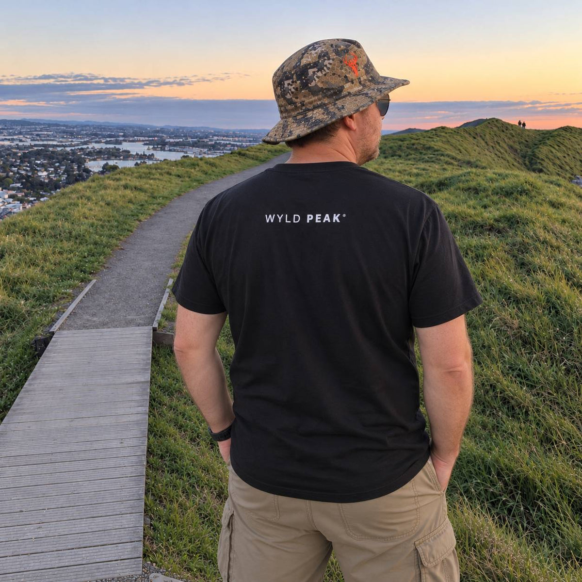 A man standing on a grassy hilltop at sunset, viewed from behind, wearing a black t-shirt with 'WYLD PEAK' printed in white on the back, khaki cargo pants, and a camouflage bucket hat. A wooden boardwalk path leads down the hill toward a city skyline.