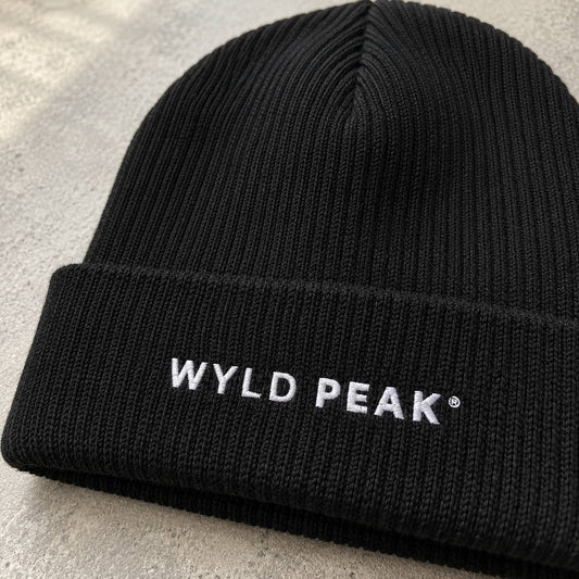 A close-up of a black ribbed knit beanie hat with 'WYLD PEAK' embroidered in white on the folded cuff, set against a light gray textured background.